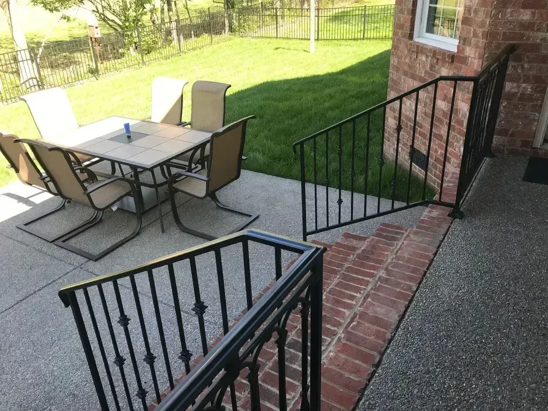 Brick patio with outdoor table, chairs, and black metal railings beside a house and grassy yard