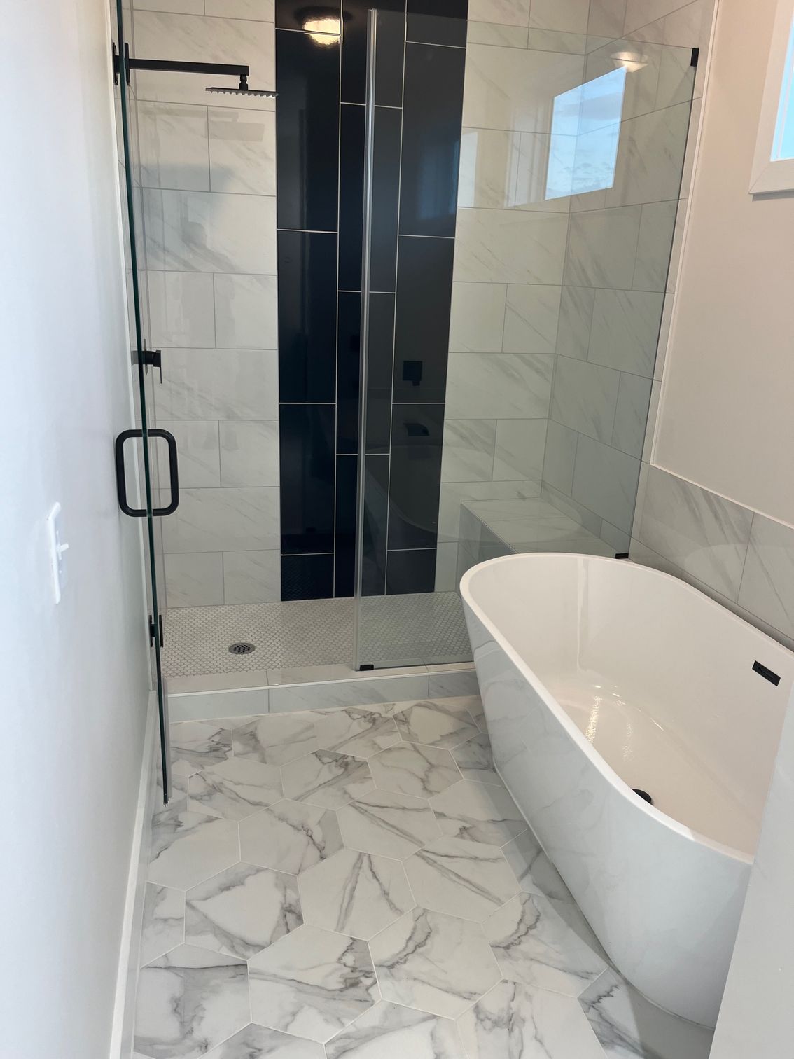 Modern bathroom with glass shower, black tile accent, marble floor, and freestanding tub