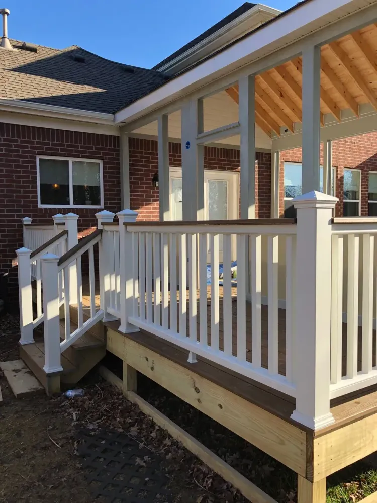 Front porch with white railing and steps attached to a brick house, viewed in sunlight