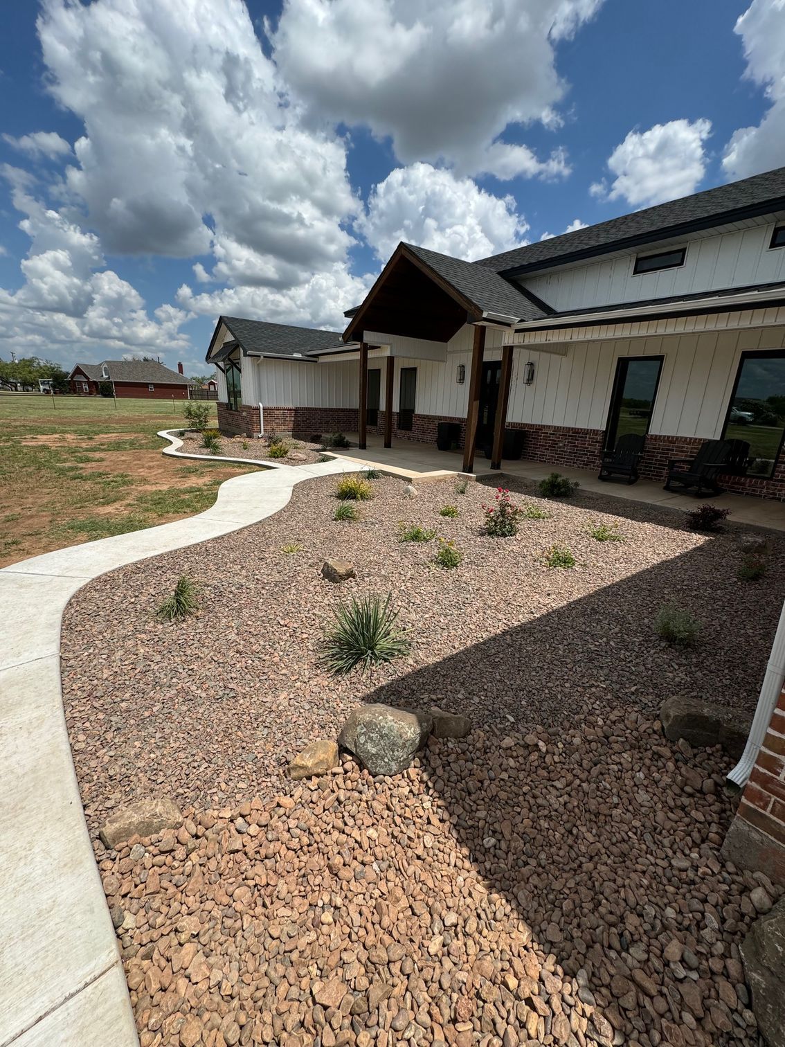 A modern white house with brick accents and a dark roof, featuring a curved concrete walkway through a rock-filled yard.