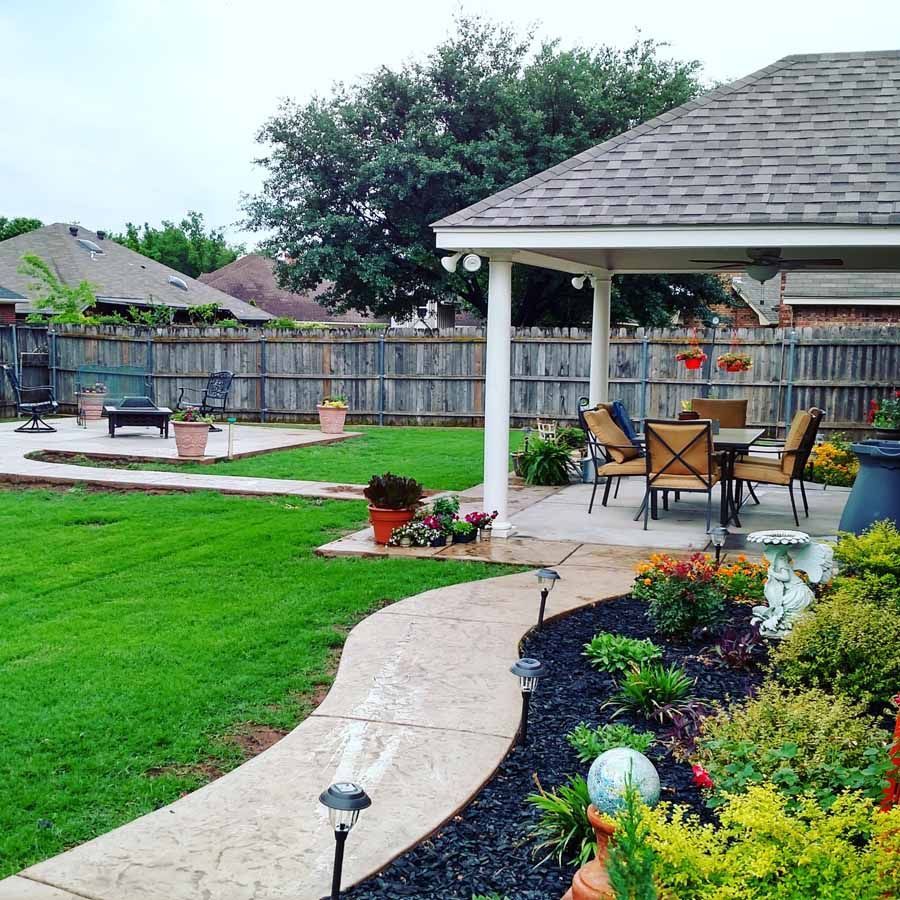 A backyard with a winding concrete path, lush green lawn, covered patio with dining furniture, and landscaped garden beds.