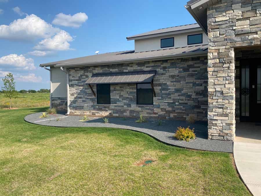 A side view of a modern house with stacked stone siding, metal roof awnings, and a curved gravel landscaping border.
