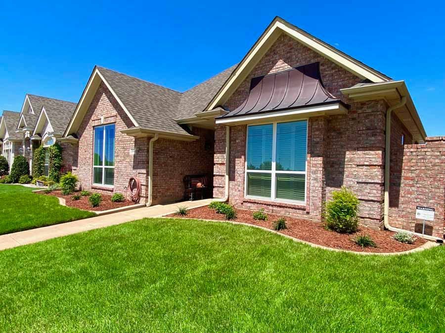 A suburban brick home with a green lawn, dark shingled roof, and arched copper-toned accent roof over a large window.