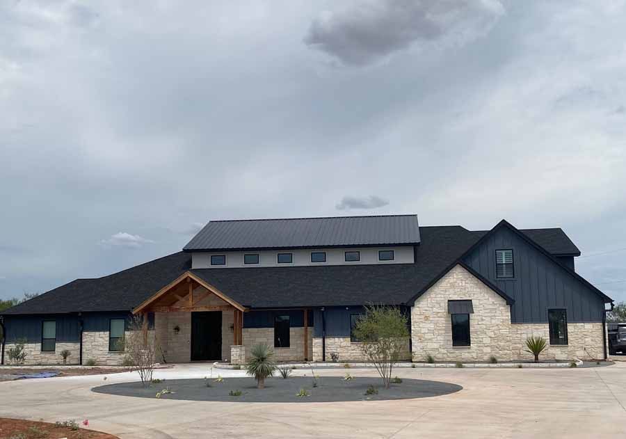 A modern farmhouse with black metal roof, vertical blue-grey siding, light stone facade, and a covered wood entrance.