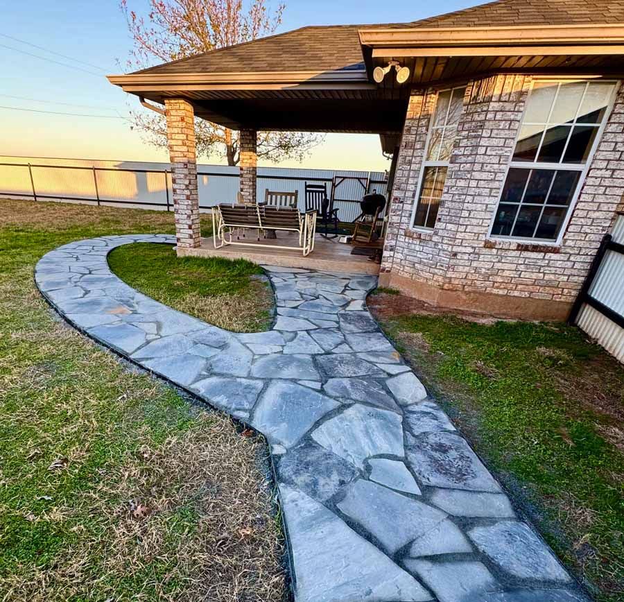 A stone walkway leads to a covered patio with a wooden swing and a brick house exterior.