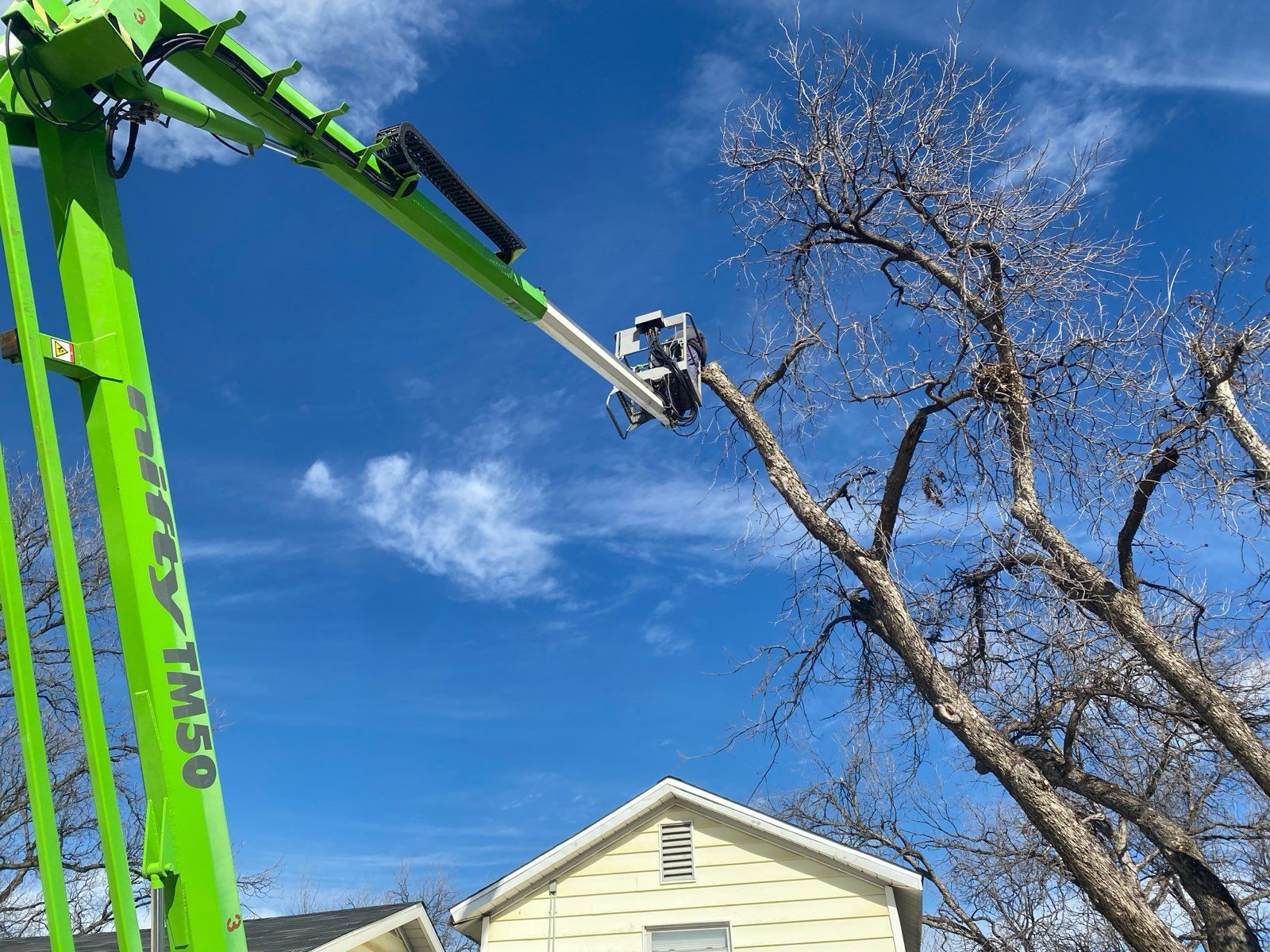 A bright green cherry picker lift extends toward the branches of a bare tree above a yellow house against a blue sky.