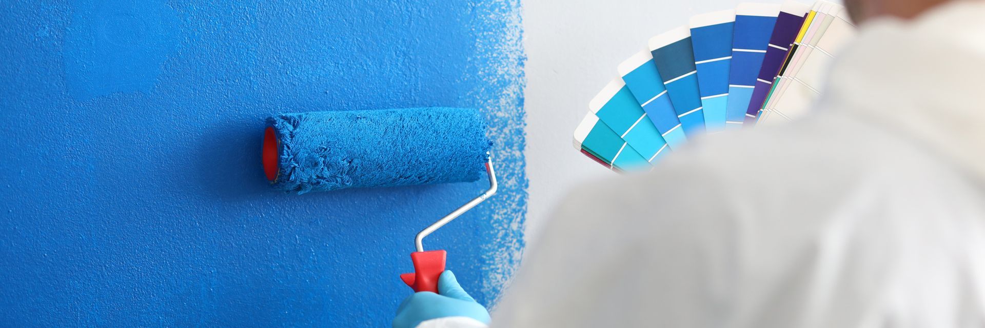 Craftsman holds a roller and a color palette and paints a white wall blue Craftsman holds a roller and a color palette and paints a white wall blue