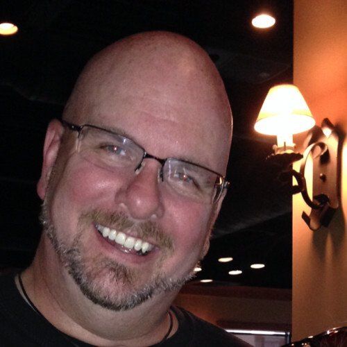 Bald man with glasses smiles at the camera, indoors, with a wall sconce lamp in the background.