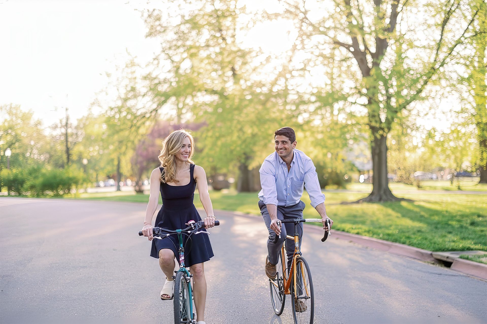 Two people ride bicycles side by side on a sunlit park road.