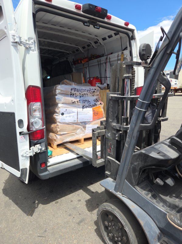Forklift unloading stacked boxed goods from the back of a white delivery van.