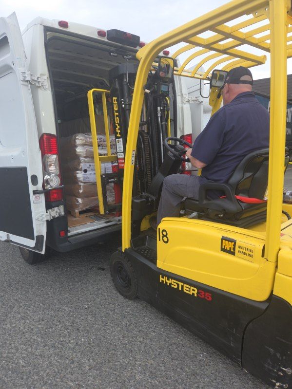 Worker operating a yellow forklift beside an open white van, loading equipment.