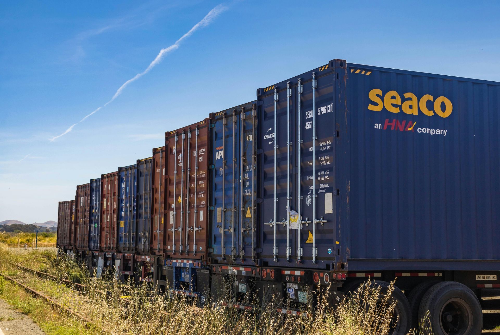 Line of shipping containers on rail cars in a grassy field under a blue sky