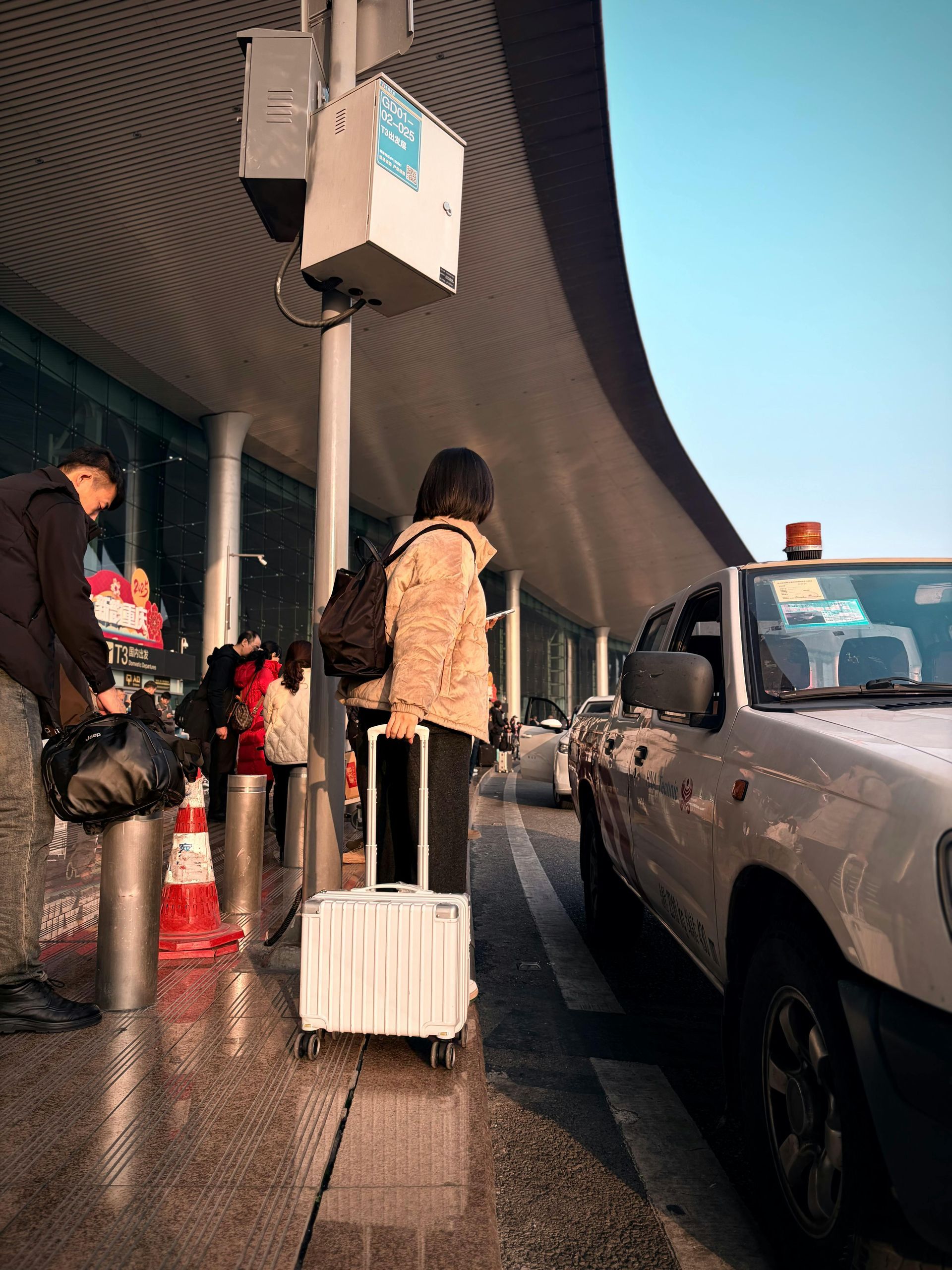 Traveler with rolling suitcase waits curbside beside a white pickup at an airport drop-off lane.