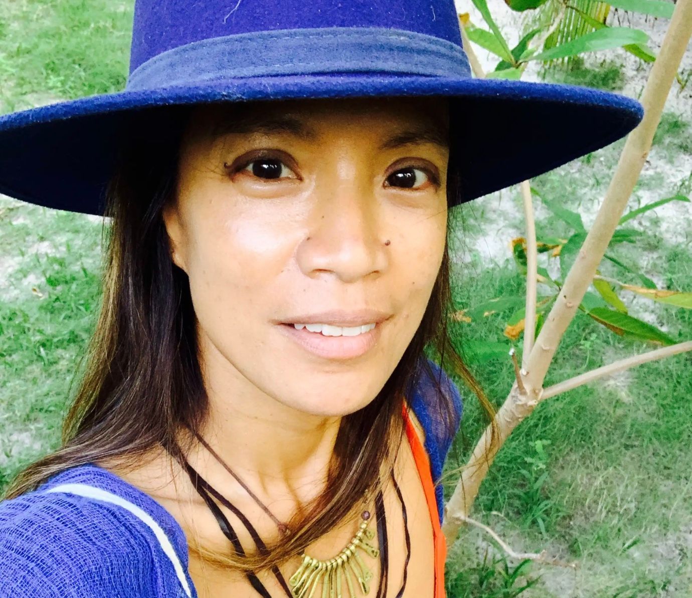 Cecilia Pagkalinawan  in blue hat smiles outdoors, near tree.