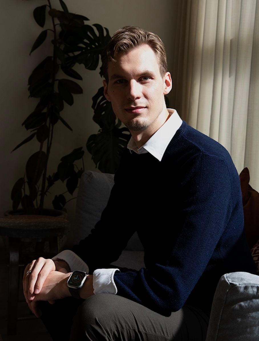 Sebastian Grube  in navy sweater and white collared shirt, looking at camera, seated near plant.