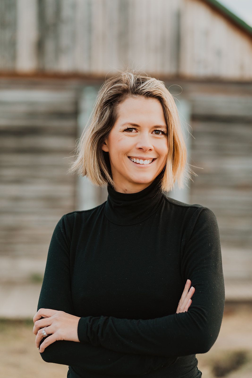 Gesche Haas  with short blonde hair smiles, arms crossed, wearing a black turtleneck. In front of a weathered wooden building.