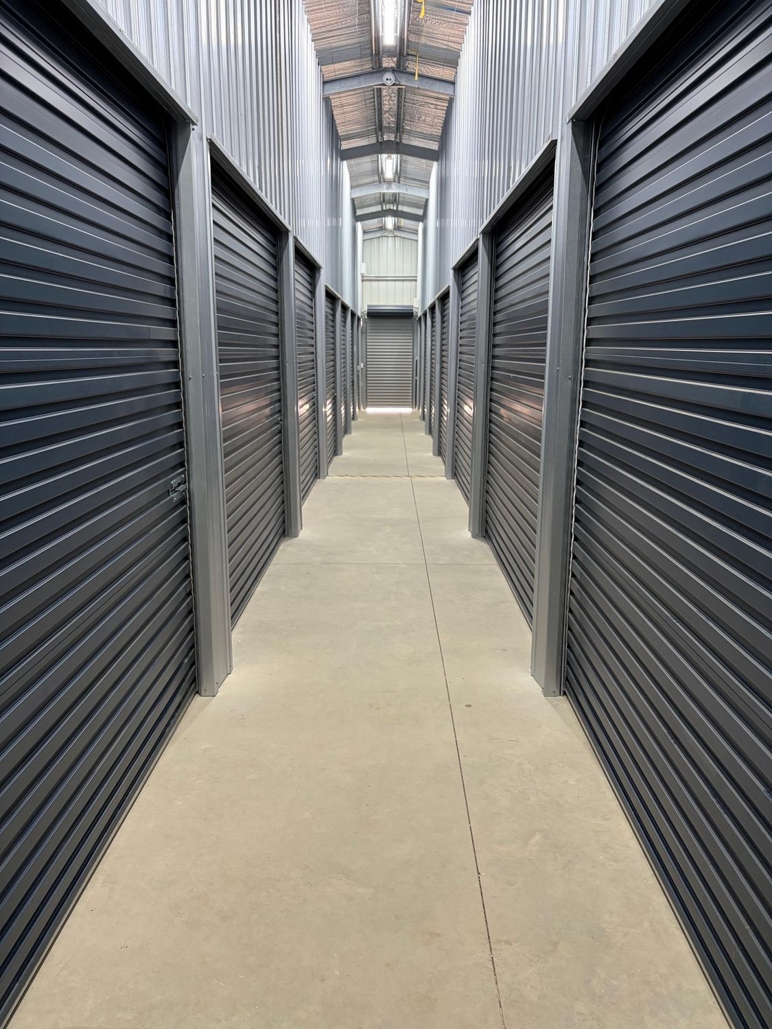 Narrow storage hallway with black roll-up doors and a concrete floor under a skylit roof