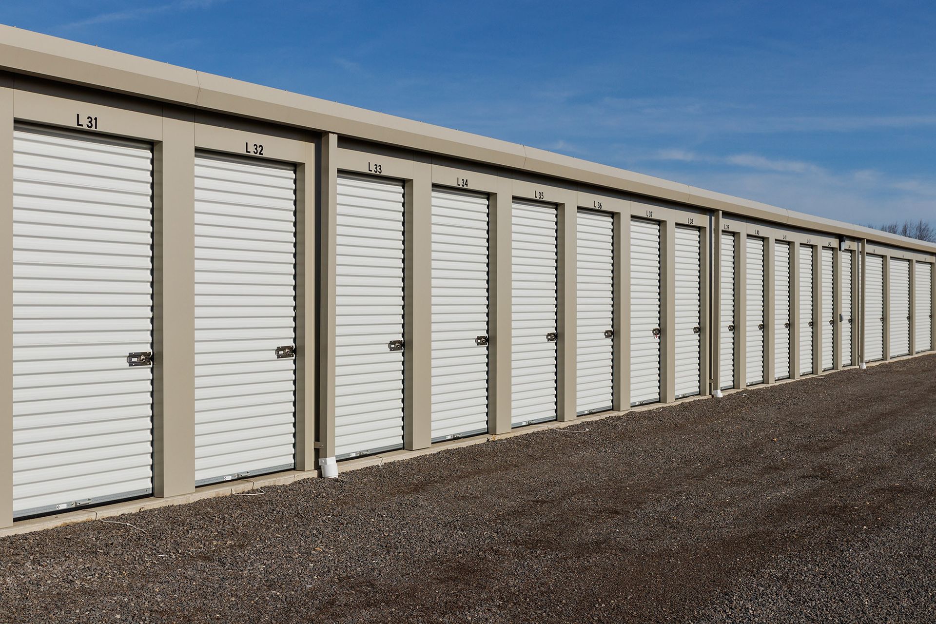Row of white storage unit doors along a beige building beside a gravel lot under a blue sky
