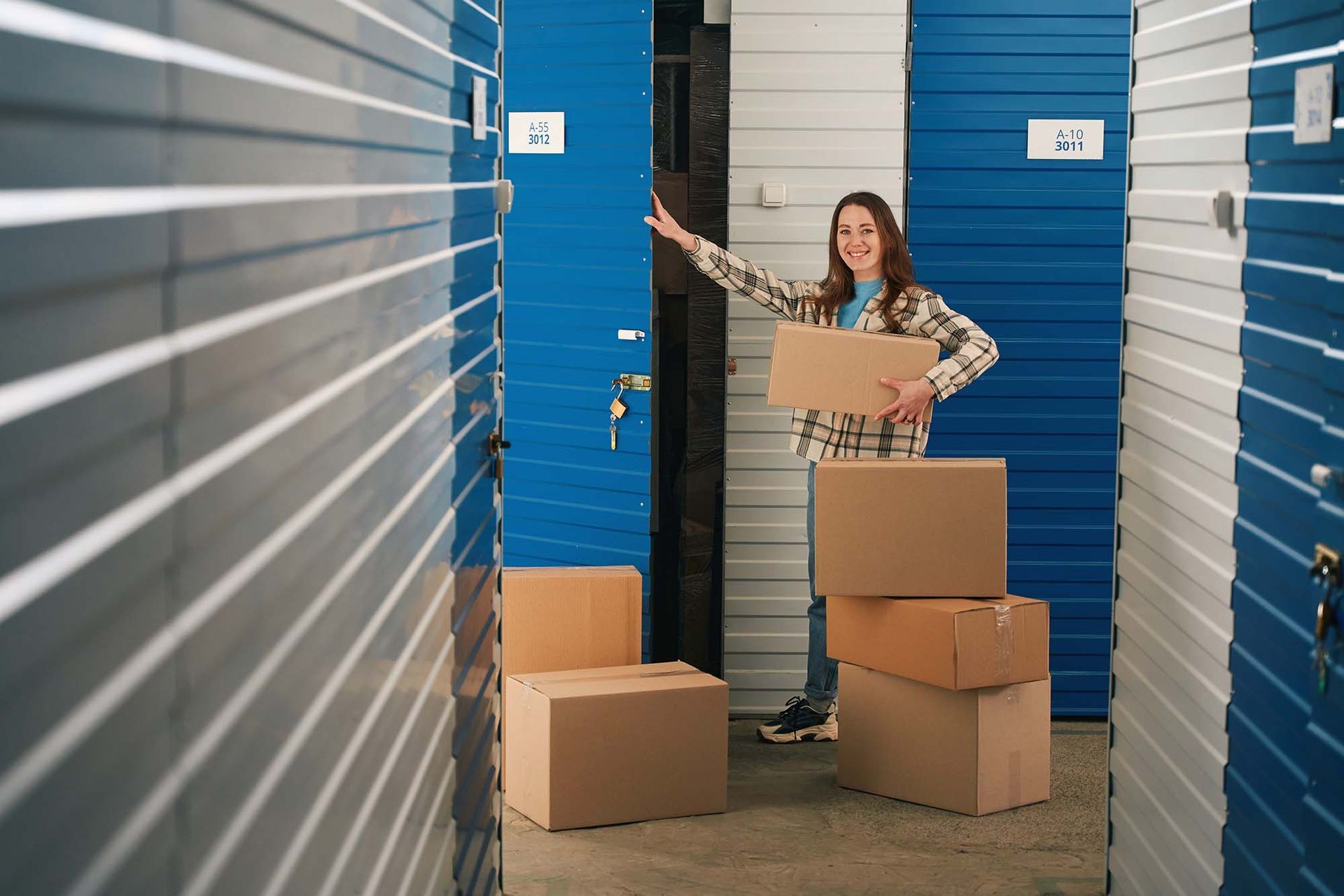 Woman stacking cardboard boxes in a storage unit aisle, smiling and reaching toward a blue door