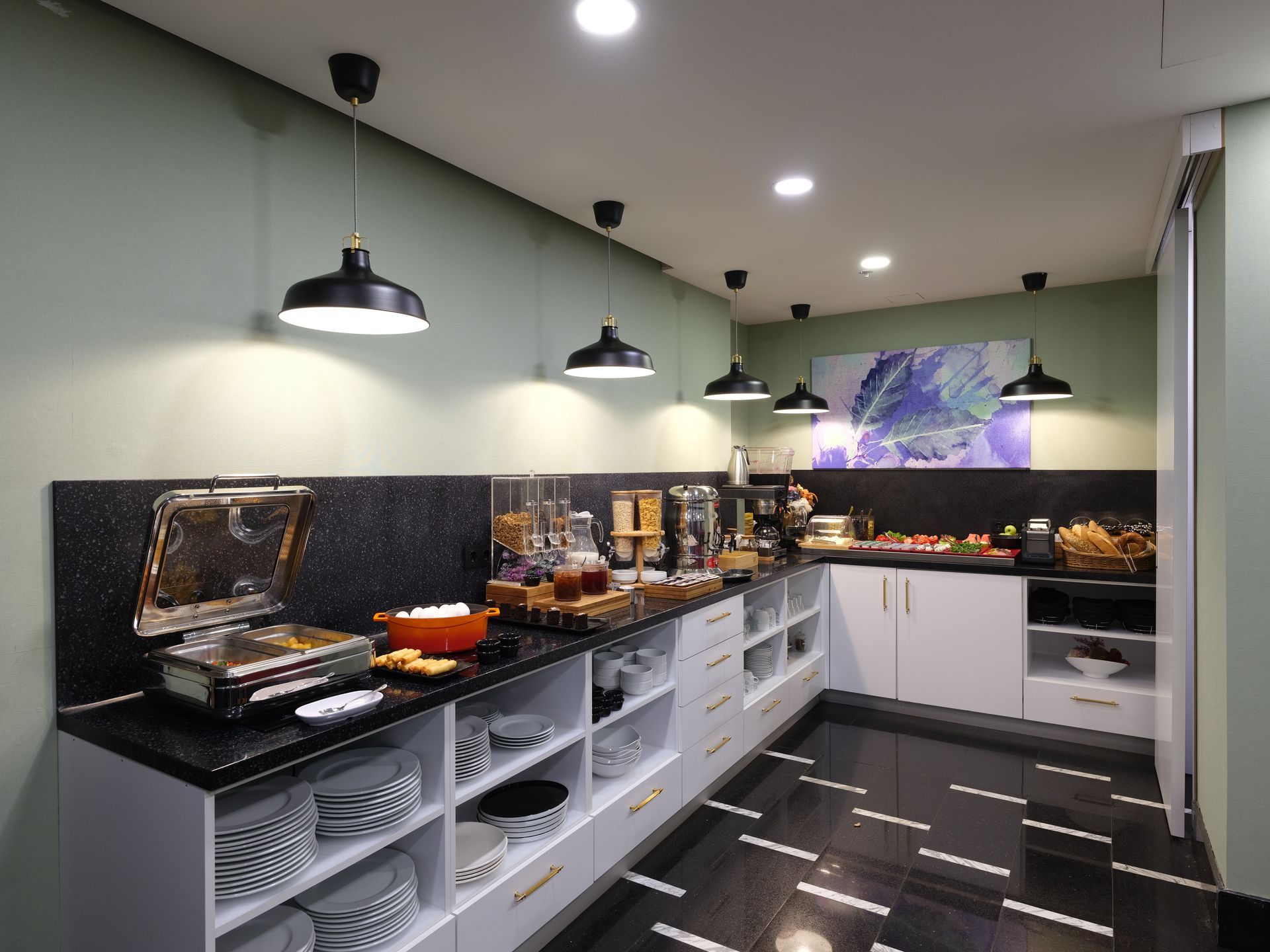 Breakfast buffet with food items on a long countertop, overhead lighting, and black and white tiled floor.