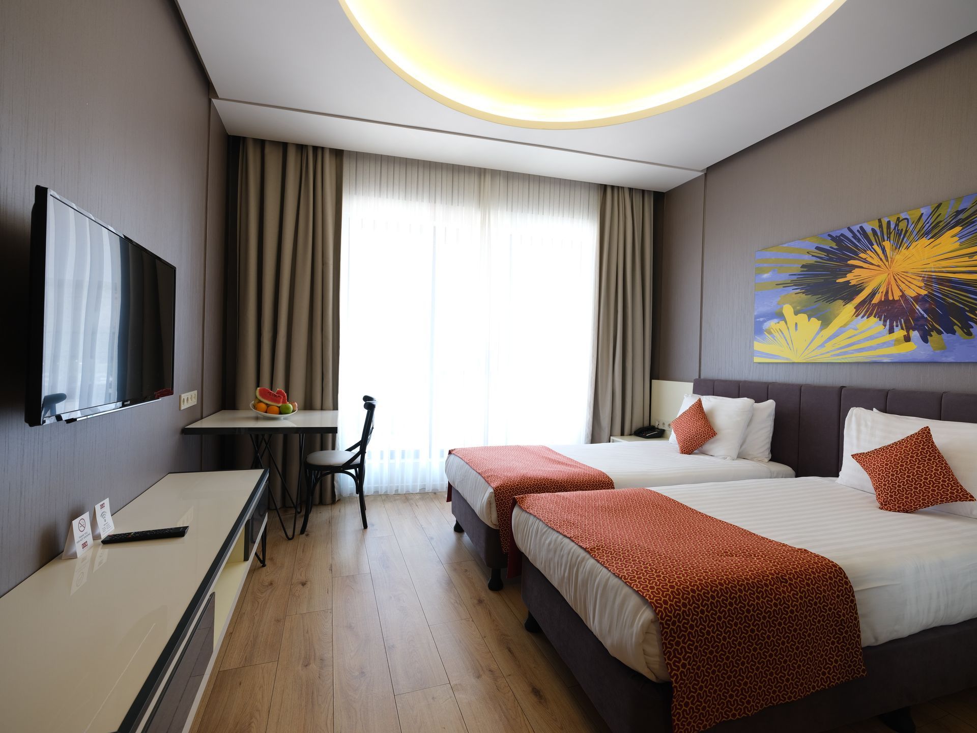 Hotel room with two twin beds, a wardrobe, artwork, and a door. The color scheme is neutral with pops of orange and blue.