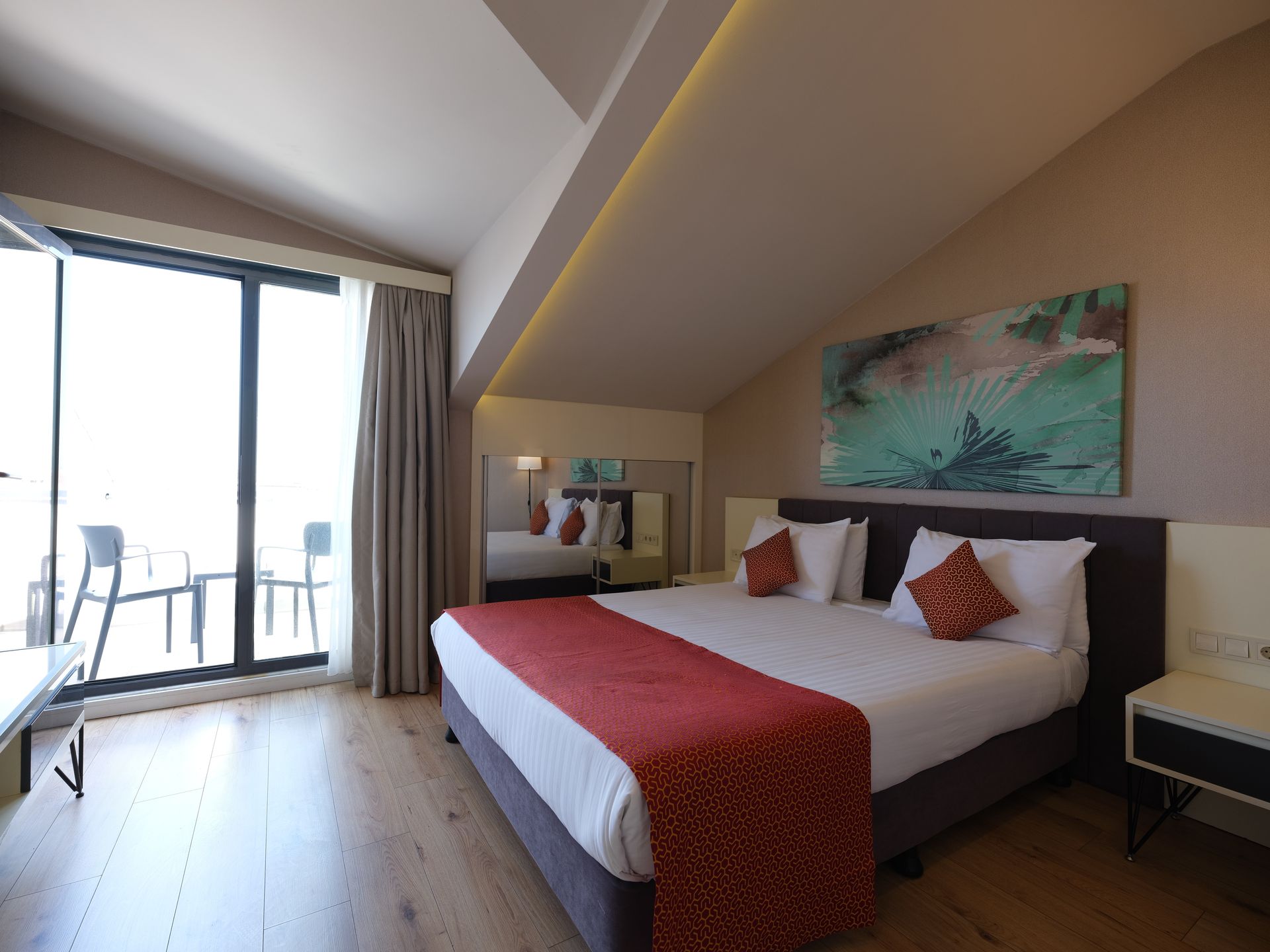 The room has light wood flooring, a large bed with red accents, and a balcony with outdoor furniture.