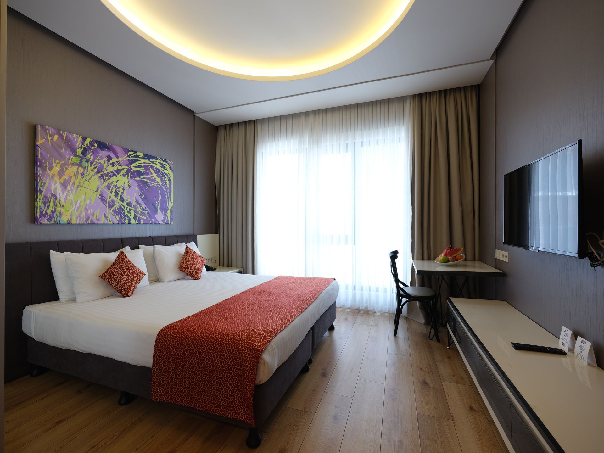 Hotel room with a king-sized bed, artwork, desk, TV, and a large window with curtains.