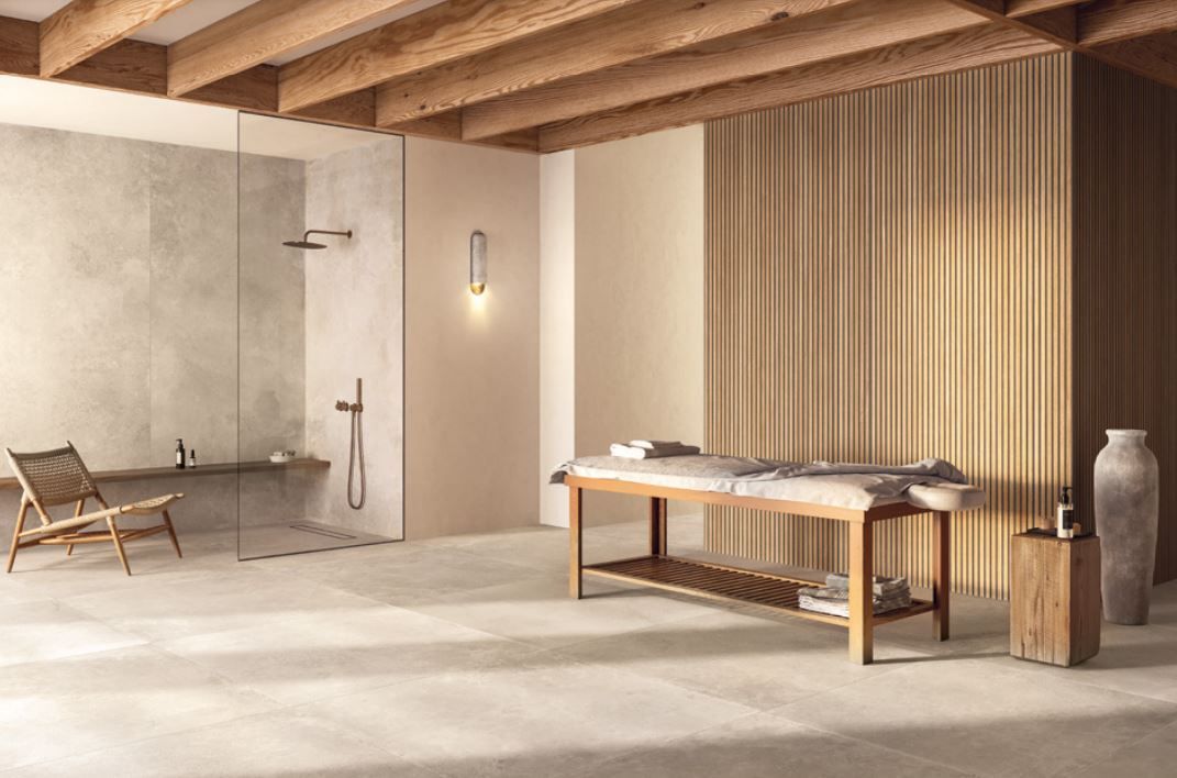 Una sala spa minimalista con lettino da massaggio, travi in ​​legno, cabina doccia e poltrona, in tonalità neutre e naturali.