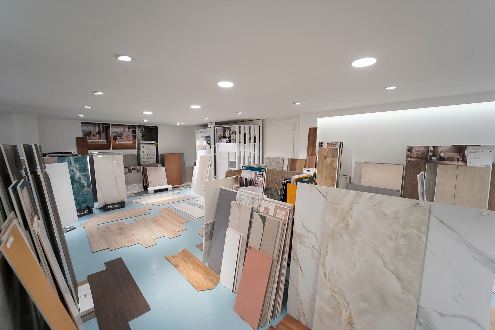 Uno showroom di piastrelle pieno di vari campioni per pavimenti e rivestimenti, disposti su scaffali e sul pavimento sotto luminose luci a soffitto.
