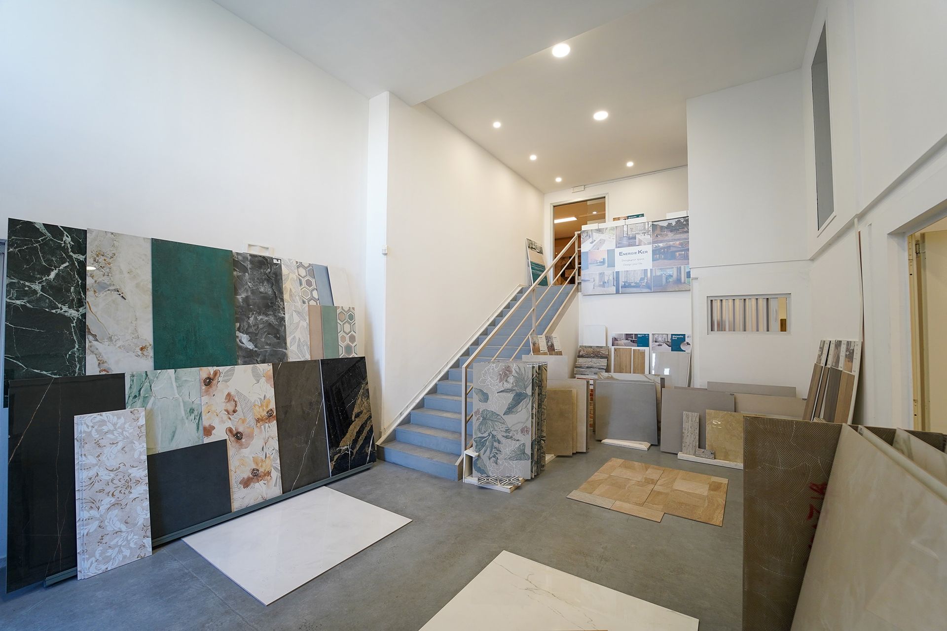 Uno showroom di piastrelle con un'esposizione di diverse lastre di pietra e marmo, una scala centrale e pavimenti dai colori neutri.