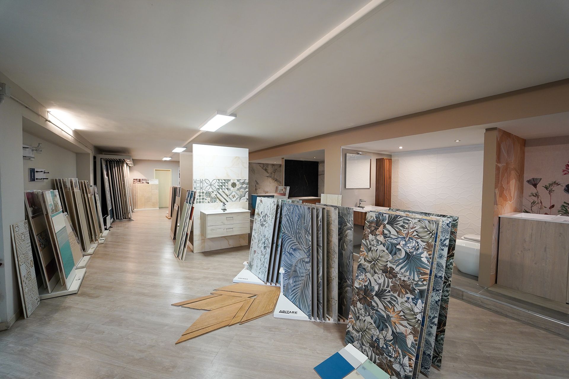 Interno di uno showroom che espone vari campioni di piastrelle in ceramica e carta da parati su degli scaffali, con pavimento in legno chiaro.