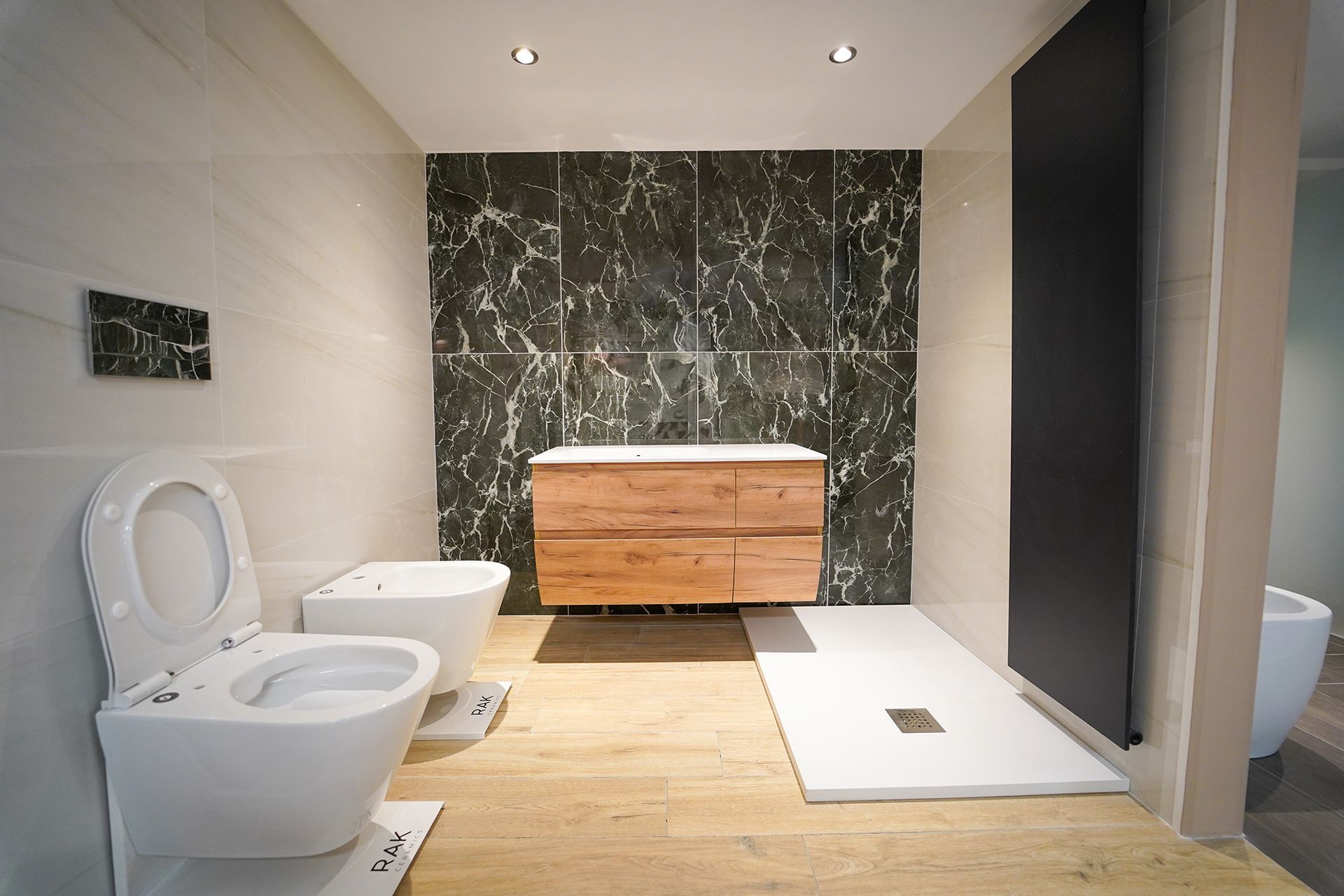 Un moderno allestimento per il bagno con WC, bidet, mobile lavabo in legno, piastrelle a parete effetto marmo e piatto doccia bianco.