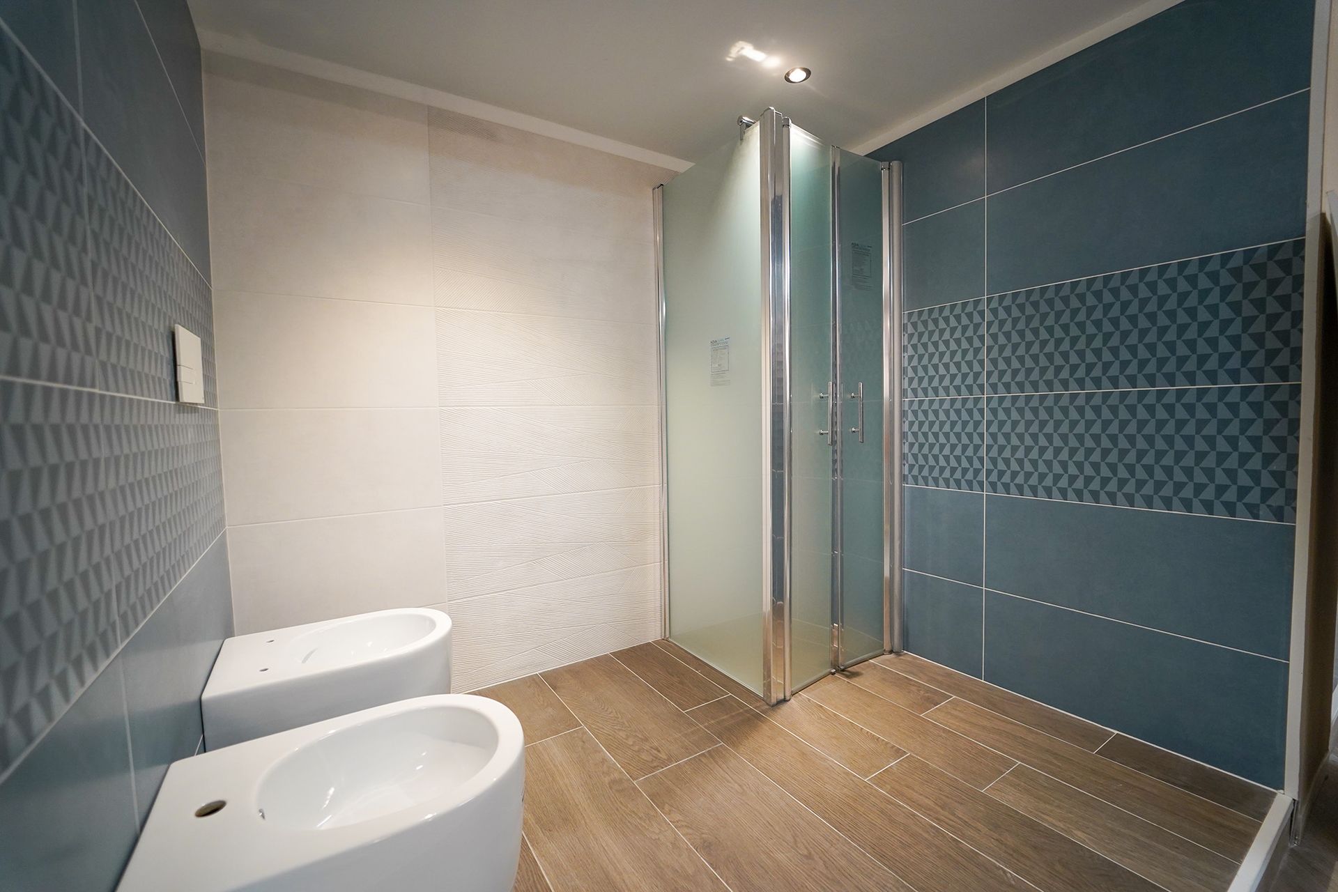 Bagno moderno con pareti rivestite con piastrelle geometriche blu e bianche, box doccia in vetro e due bidet in porcellana bianca.