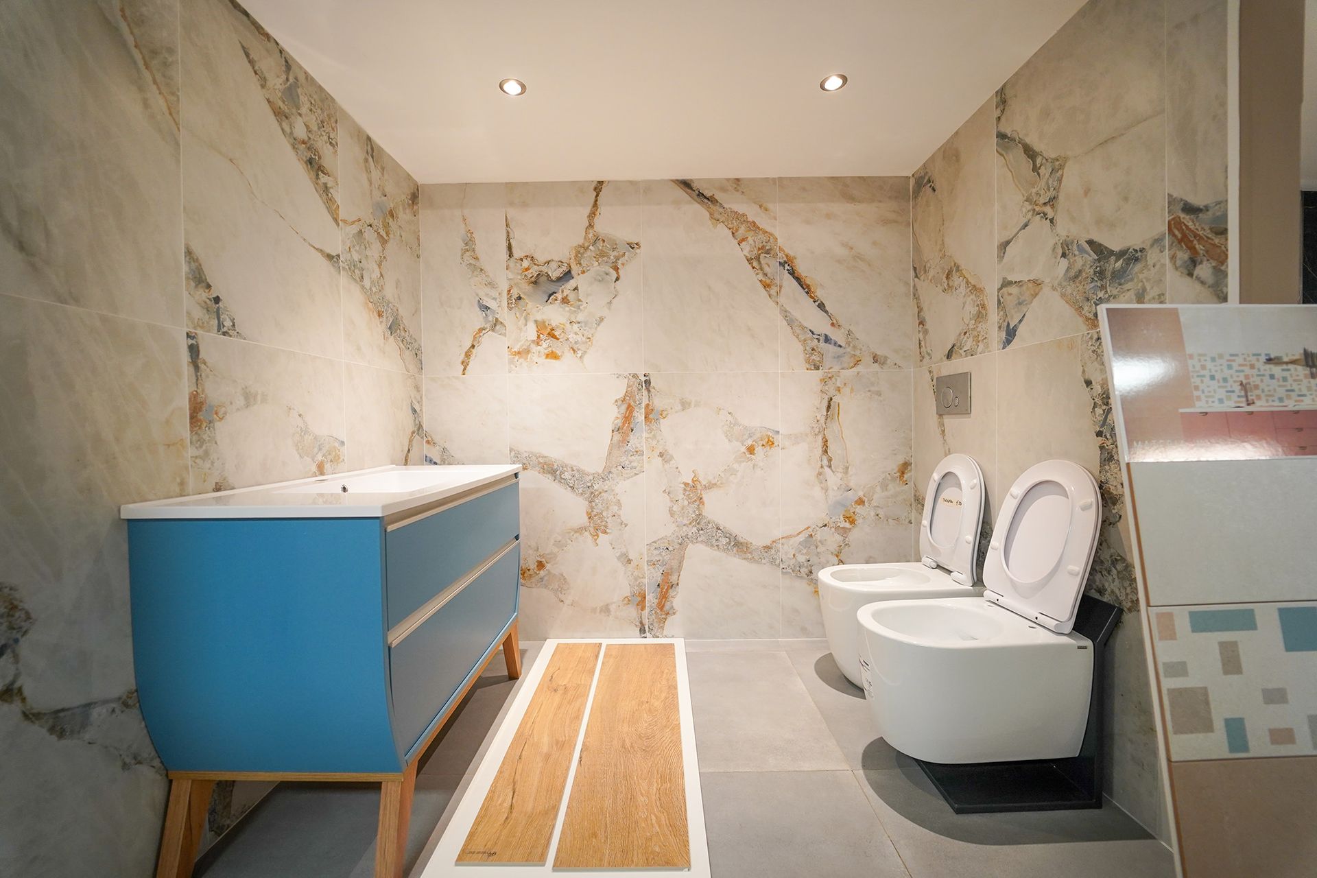 Un bagno moderno con mobile lavabo blu, pavimento con inserti effetto legno e due WC bianchi su pareti con motivo marmorizzato.