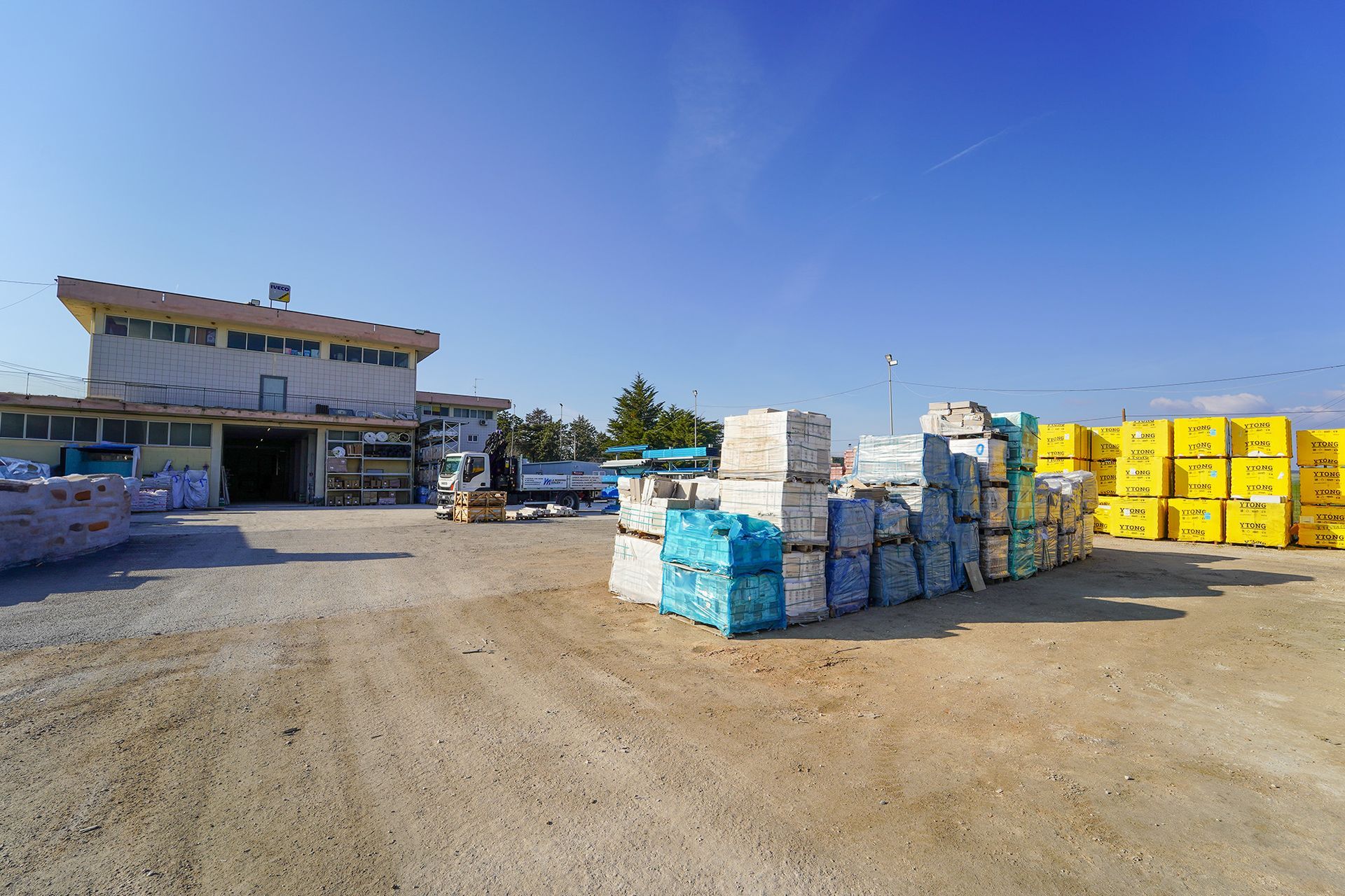 Un'inquadratura ampia di un edificio con pile di pallet imballati, alcuni blu e altri giallo brillante, in un polveroso deposito all'aperto.