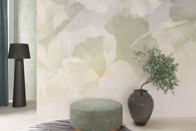 Una stanza con un murale a motivi di foglie, un pouf verde salvia, un vaso scuro con una piccola pianta e una lampada da terra alta.