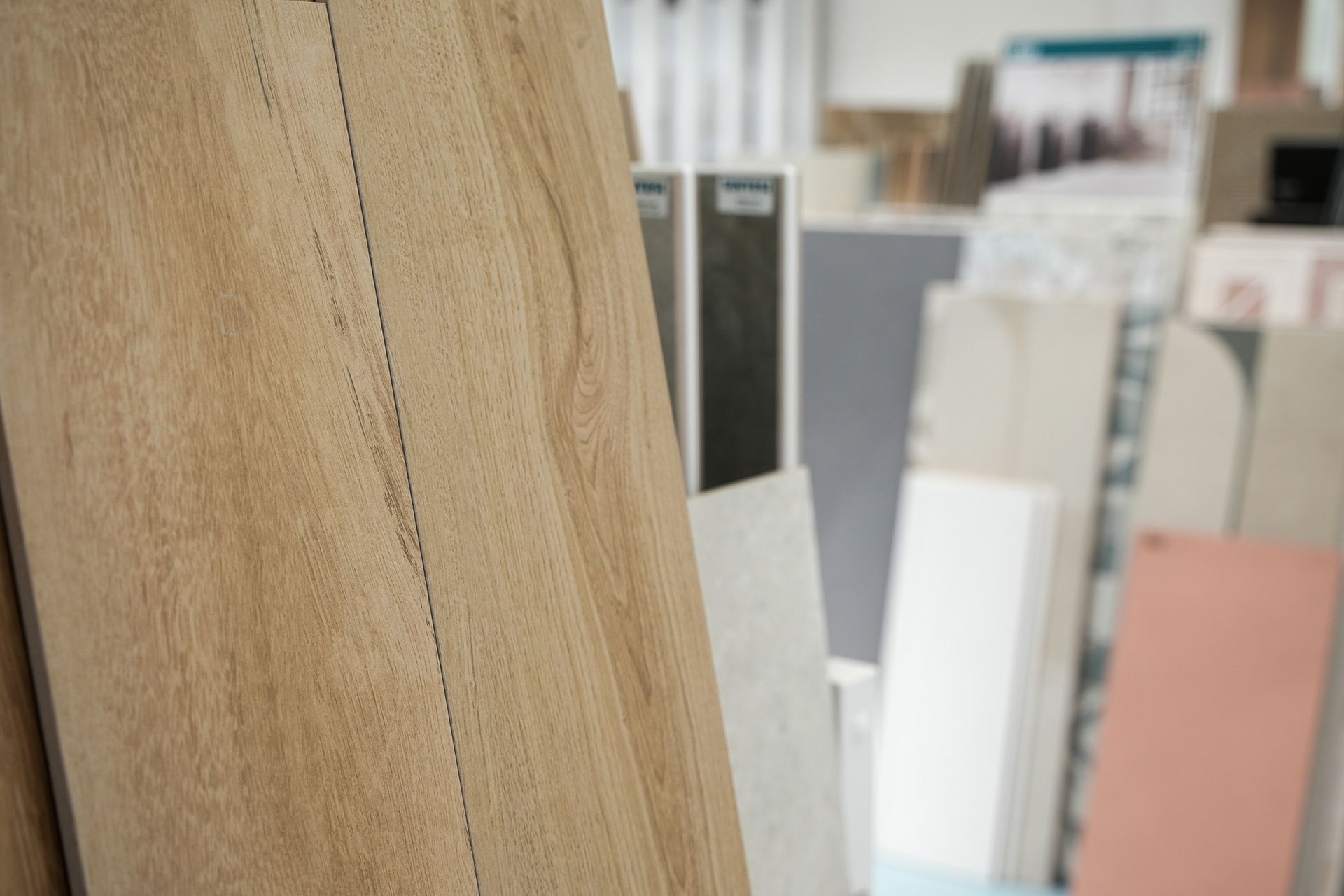 Primo piano di una piastrella per pavimento effetto legno chiaro in uno showroom, con vari altri campioni di piastrelle sullo sfondo sfocato.