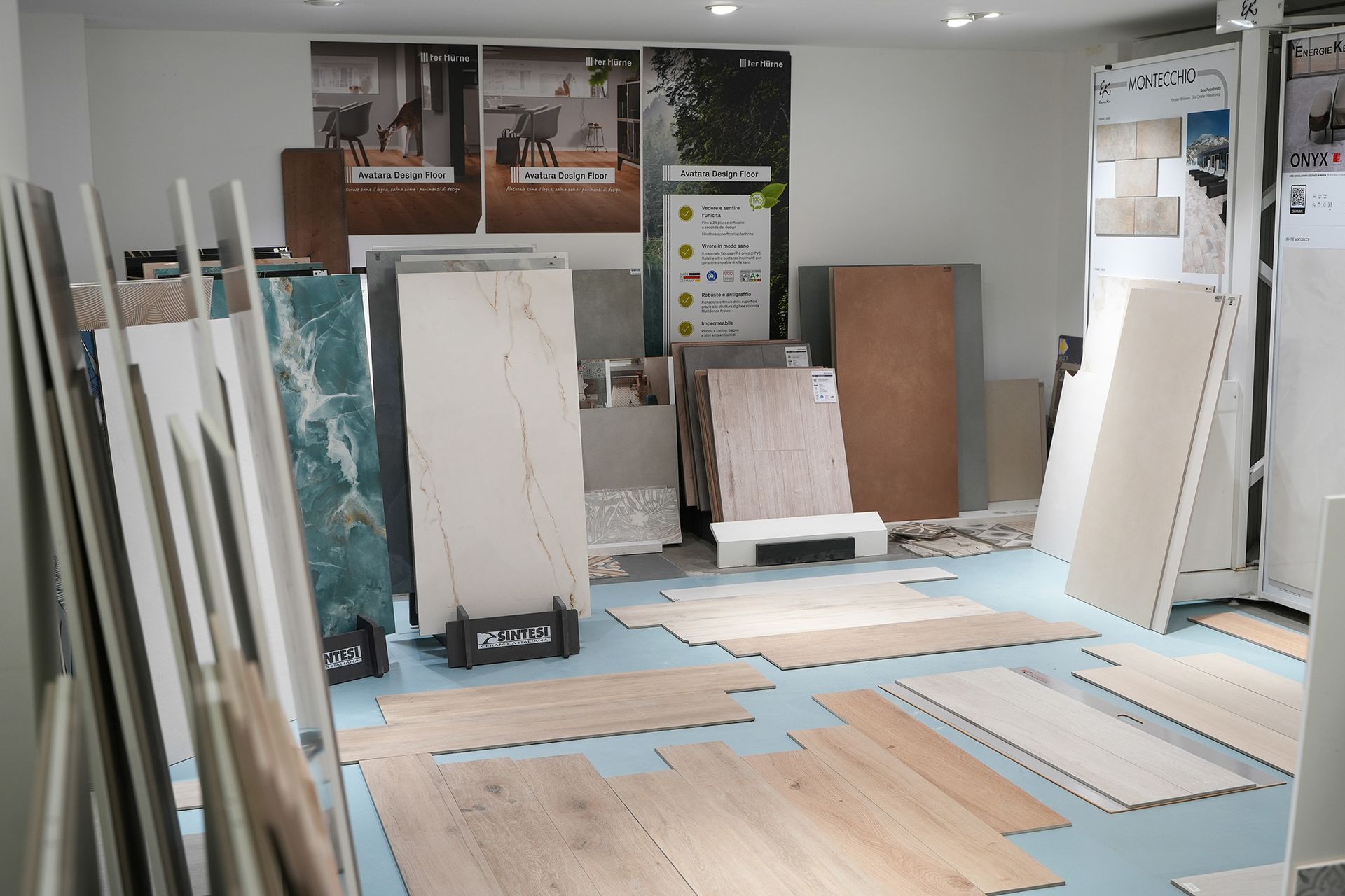 Uno showroom espone vari campioni di piastrelle e pavimenti in legno, con pannelli verticali in piedi e pezzi posati sul pavimento.
