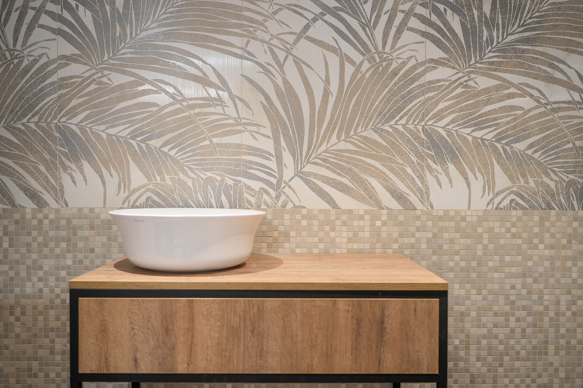 Un lavabo a ciotola bianco è posizionato su un mobiletto in legno, sullo sfondo di piastrelle a mosaico e piastrelle a parete con motivi di foglie tropicali.