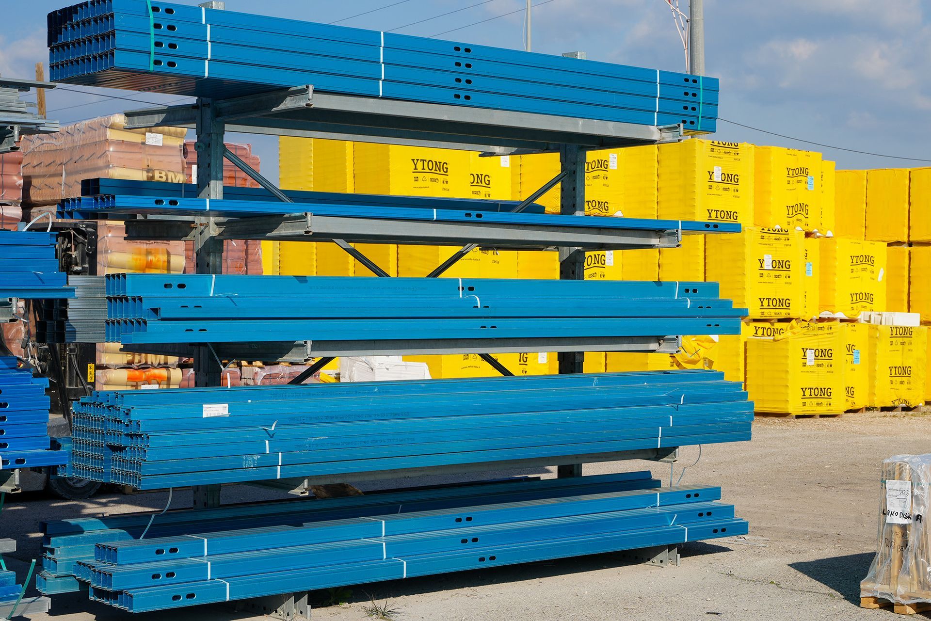 All'aperto, una scaffalatura che sorregge travi metalliche blu impilate, con contenitori gialli sullo sfondo.