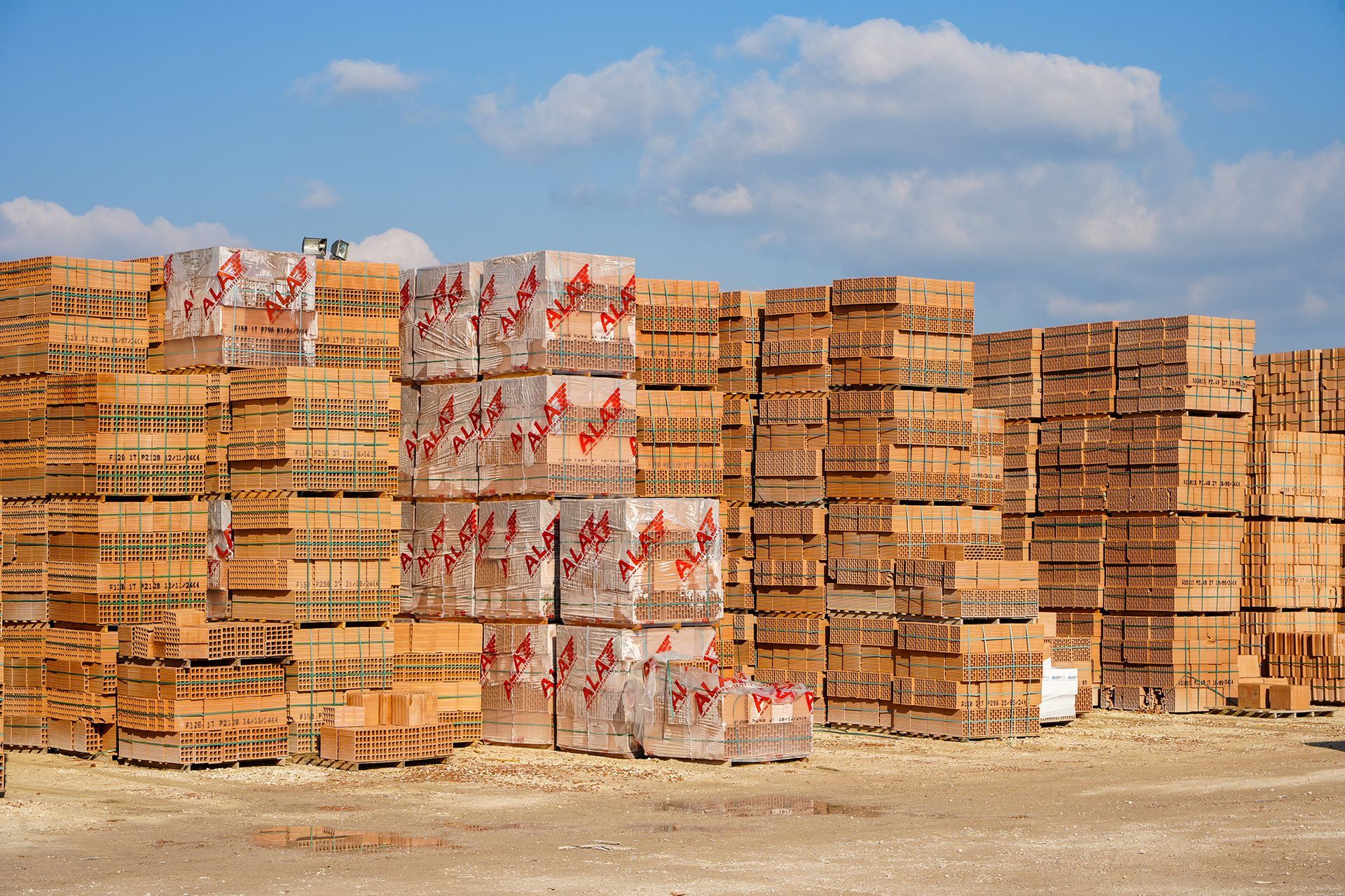 Pallet impilati di mattoni rosso-marroni, stoccati all'aperto in un terreno sterrato sotto un cielo azzurro.