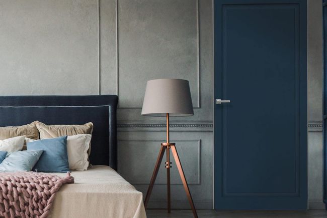 Una camera da letto con testiera blu scuro, lampada da terra a treppiede e porta blu scuro abbinata, su una parete rivestita di pannelli grigi.