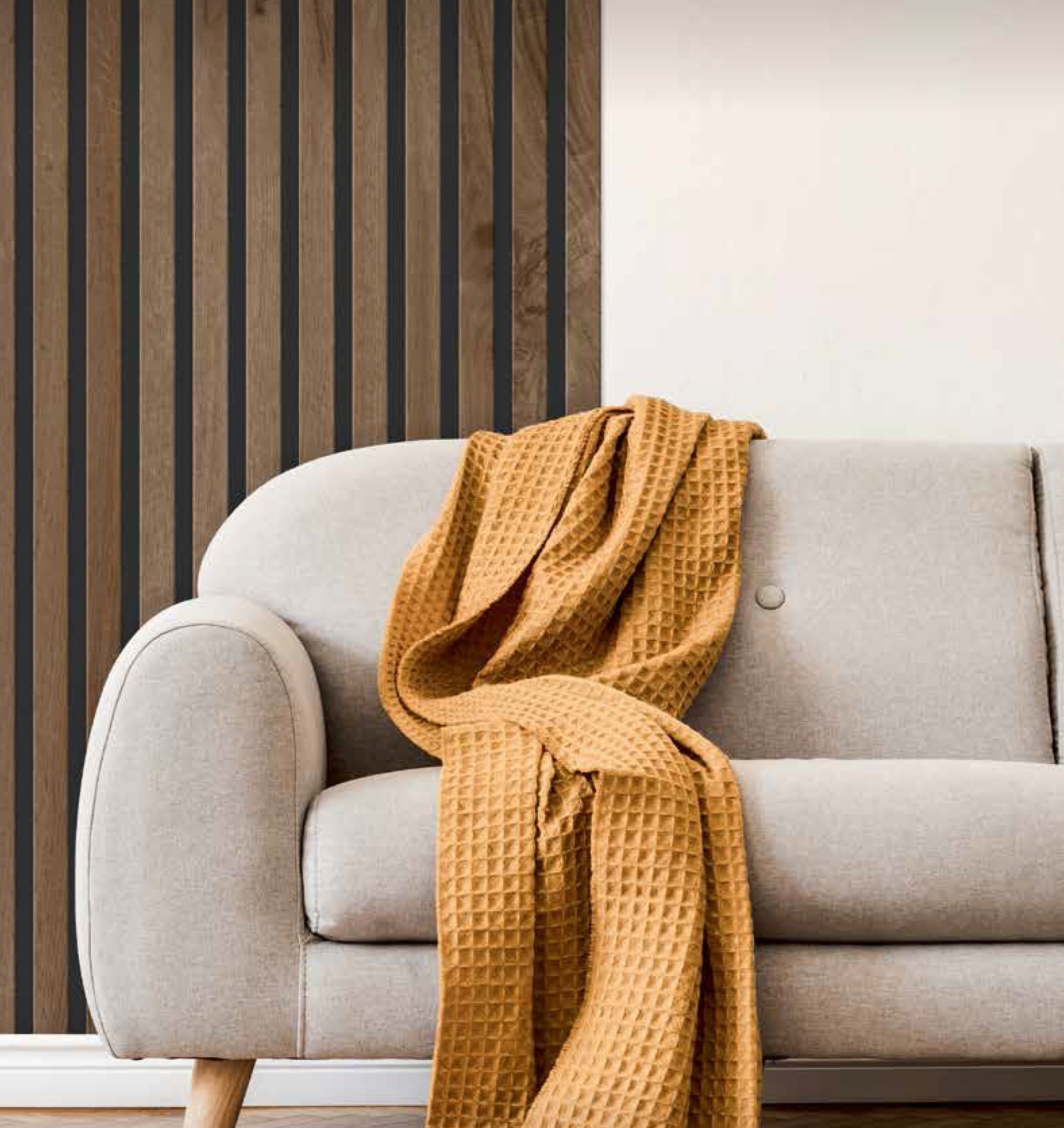 Un divano color beige chiaro con una coperta a nido d'ape giallo oro drappeggiata sopra, appoggiato a una parete con listelli di legno verticali.