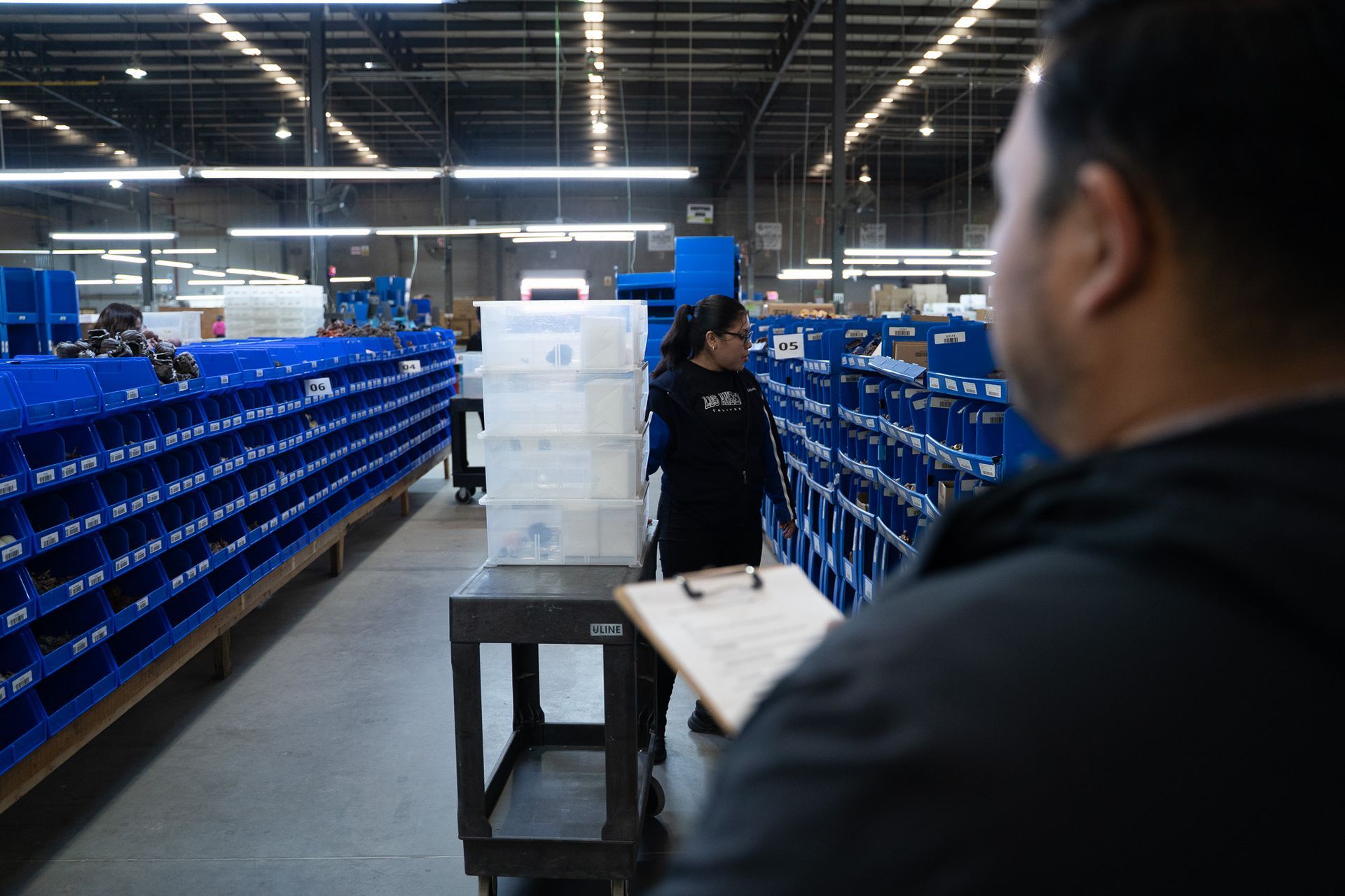 Warehouse aisle with blue bins and workers moving stacked white containers on a cart