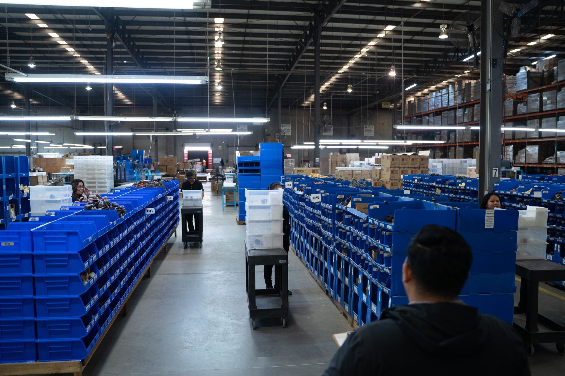Warehouse aisle with rows of blue bins and workers under bright industrial lights