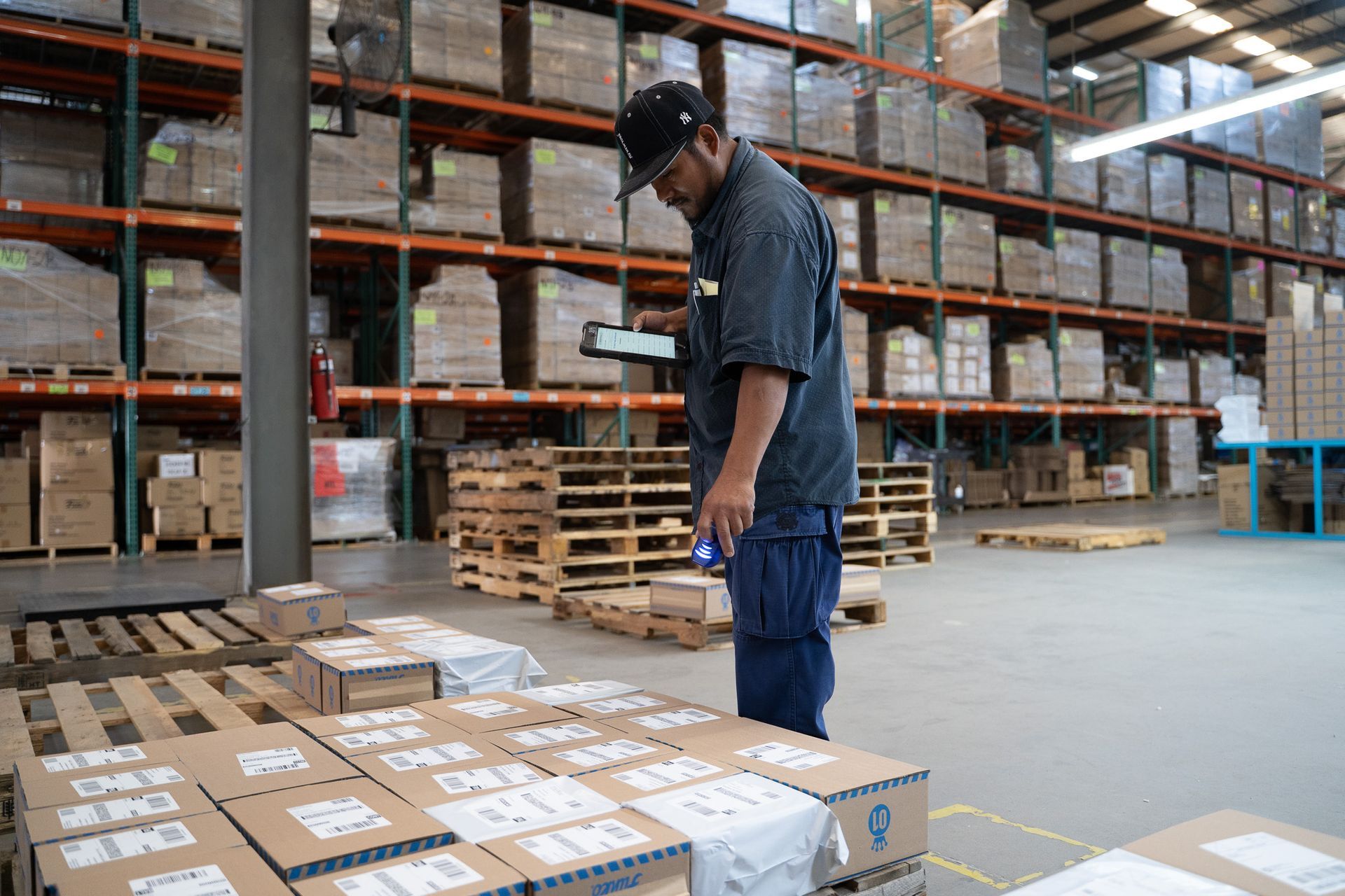 Worker standing among stacked boxes in a warehouse aisle, checking a package on a pallet.