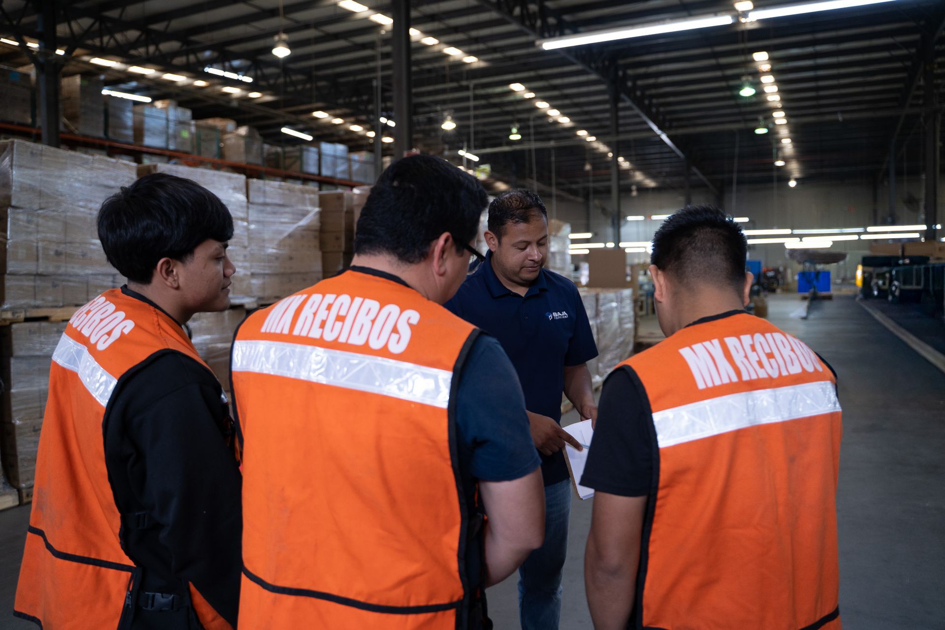 Three workers in orange safety vests stand in a warehouse, looking at a man in a dark shirt.