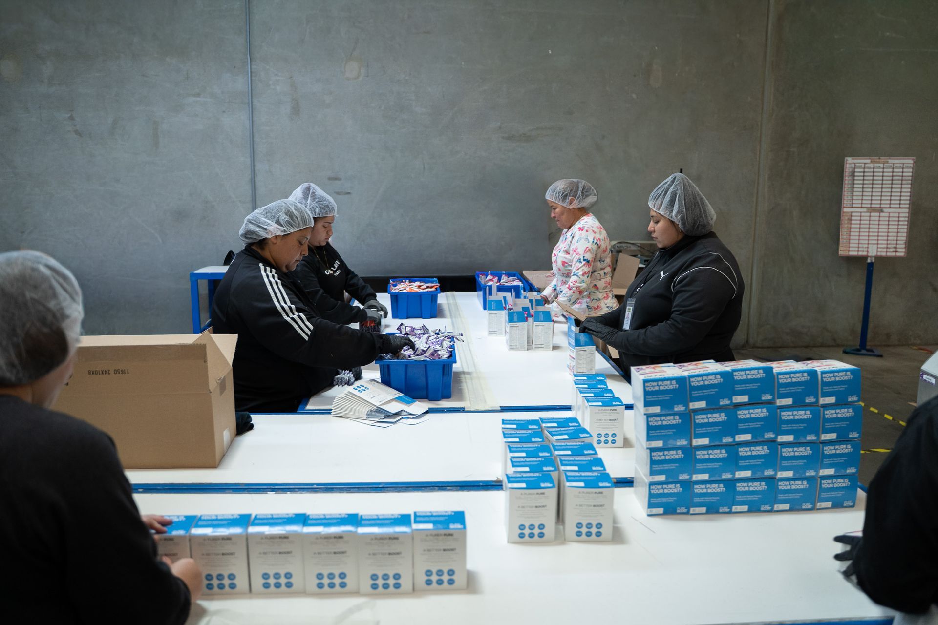 Workers in hairnets packing blue boxes at a long table in a warehouse room