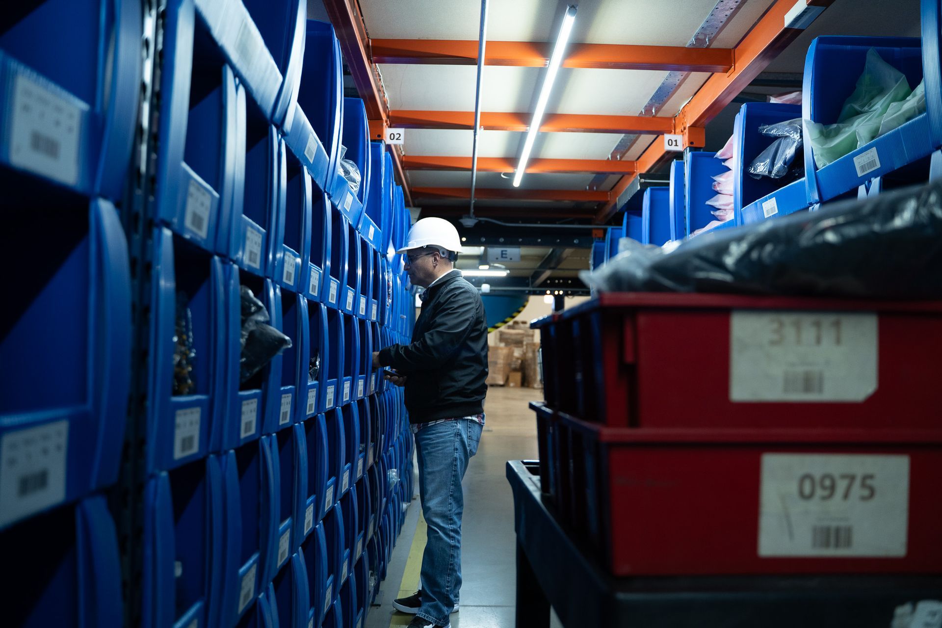 Worker in white hard hat using a tablet in a warehouse aisle lined with blue storage bins