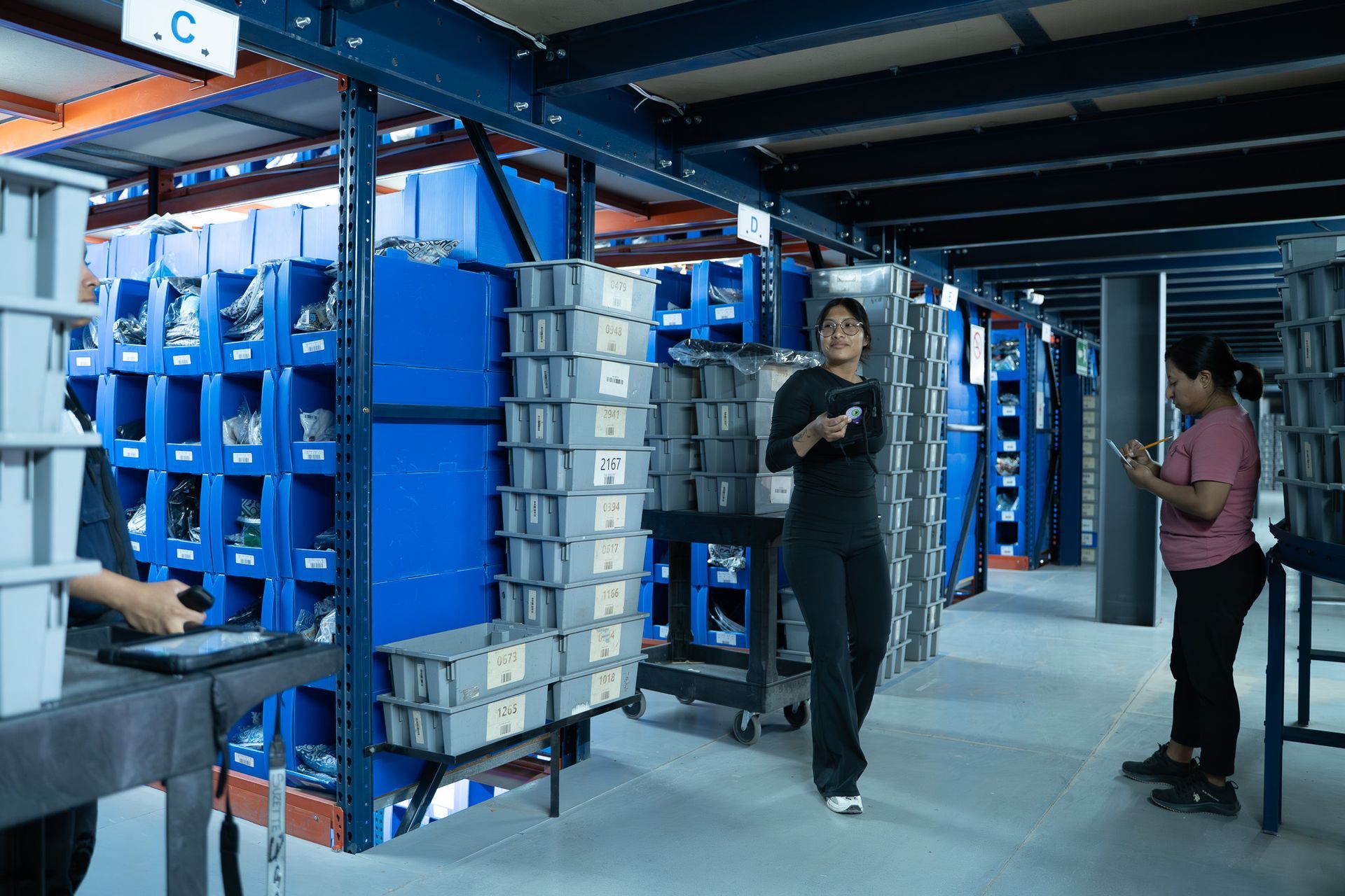 Warehouse aisle with blue shelves and stacked boxes; two people stand and walk nearby.